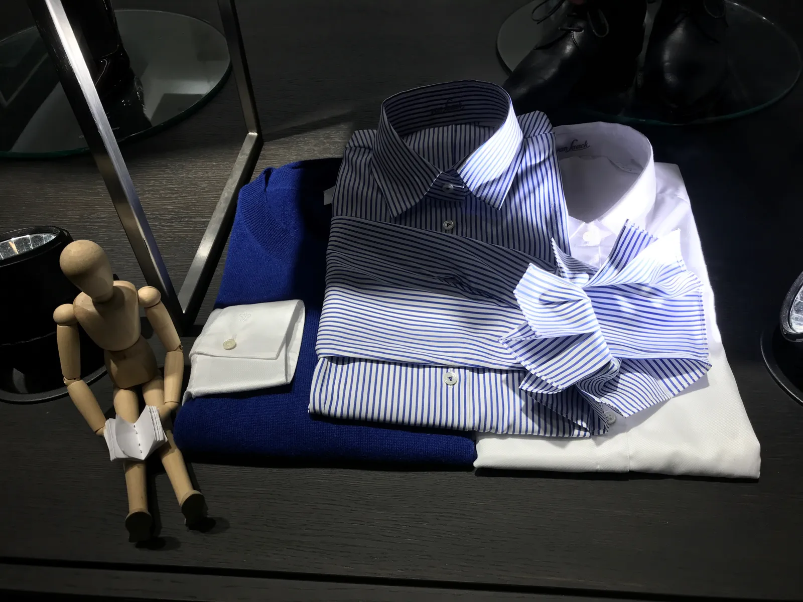 Bodegón en tienda con camisas de rayas dobladas, un jersey azul y un maniquí articulado de madera sentado al lado